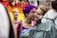 Two individuals share a close, intimate moment in a colorful, public setting. One person has vibrant purple hair and wears glasses, while the other is smiling warmly. In the background, a rainbow flag suggests a celebration of diversity and inclusivity.