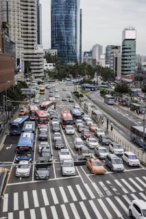 A busy urban intersection with multiple lanes of vehicles, including buses, cars, and taxis, waiting at a traffic light. Modern skyscrapers and buildings line the streets in the background. Pedestrians can be seen on sidewalks and crossing the street.