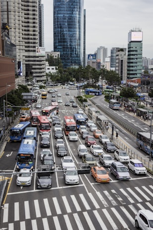 A busy urban intersection with multiple lanes of vehicles, including buses, cars, and taxis, waiting at a traffic light. Modern skyscrapers and buildings line the streets in the background. Pedestrians can be seen on sidewalks and crossing the street.
