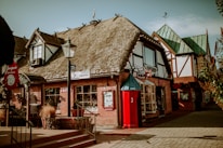 A quaint, vintage-style shop with a thatched roof and brick exterior stands in a pedestrian area. It's adorned with signs, including 'TOBAK SHOPPE' and 'VIKING GARDEN Restaurant'. A red vintage telephone booth adds a nostalgic touch. Plants and decorative elements enhance the charming atmosphere.