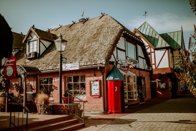 A quaint, vintage-style shop with a thatched roof and brick exterior stands in a pedestrian area. It's adorned with signs, including 'TOBAK SHOPPE' and 'VIKING GARDEN Restaurant'. A red vintage telephone booth adds a nostalgic touch. Plants and decorative elements enhance the charming atmosphere.