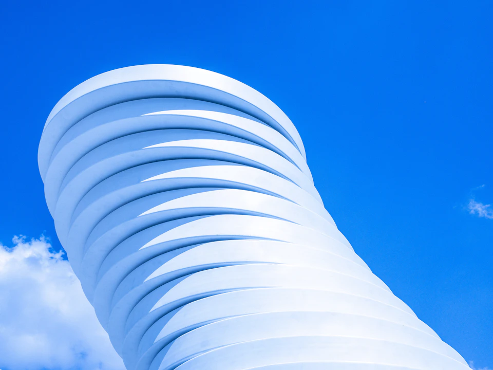 The image shows a striking white building with a unique curved design, resembling a spiral staircase or a wave-like structure. The building is set against a clear blue sky with fluffy white clouds scattered across it. The perspective of the photo is from below, looking up at the impressive height and intricate details of the architectural marvel.
