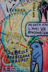 Colorful graffiti art featuring a mix of abstract and recognizable elements, including a stylized humanoid figure, sketches of faces, and various texts in different fonts and colors spread throughout. The artwork is vibrant with a mix of flowing lines and sharp outlines, incorporating a mix of languages and styles.