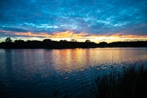 A serene lake reflects the vibrant hues of a sunset sky with a mixture of blues, oranges, and yellows. The silhouette of trees and shrubs lines the horizon, creating a peaceful and picturesque scene. The water surface is slightly rippled, capturing the colors and adding texture to the image.