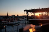 A rooftop setting at sunset with tables and chairs. The sun is setting in the distance, casting a warm glow over the city skyline, which features a prominent spire and other buildings. Parts of the rooftop are covered with a decorative trellis adorned with foliage.