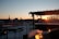 A rooftop setting at sunset with tables and chairs. The sun is setting in the distance, casting a warm glow over the city skyline, which features a prominent spire and other buildings. Parts of the rooftop are covered with a decorative trellis adorned with foliage.