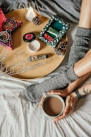 A cozy setup featuring a person with tattooed arms holding a mug of coffee, wearing gray socks. A round tray holds various holiday-themed skincare products, pine cones, a small candle, and decorative branches. The scene is set on a bed with soft gray and white linens, creating a warm and relaxing atmosphere.