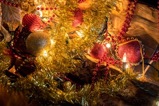 A festive arrangement featuring red and gold ornaments, tinsel, and string lights. The decorations include round baubles and beads, surrounded by sparkling garlands. Warm lighting creates a cozy and inviting atmosphere.
