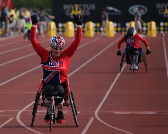 A wheelchair athlete wearing a red uniform with 'United Kingdom' printed on it raises their arms in celebration while crossing the finish line on a track. Another wheelchair athlete in similar attire is behind them. The background features people and a banner with 'Invictus Games'.