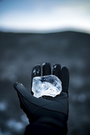A gloved hand holds a translucent piece of ice against a blurred, dark background. The ice is clear and cool, reflecting the surrounding light.