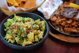 A close-up view of a black bowl filled with freshly made guacamole, consisting of diced avocado, tomatoes, onions, and cilantro. In the background, there is a basket lined with white paper containing crispy tortilla chips. Additionally, a plate with grilled meat and a wrapped foil item can be partially seen, indicating a platter of assorted Mexican dishes.