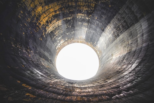 A large industrial structure resembling a cooling tower with visible concrete walls and an open circular top. The interior walls are textured with uneven patterns, and light streams in from the open top, creating a dramatic effect.