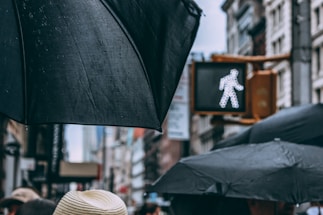 Large black umbrellas are prominently displayed against an urban backdrop. A pedestrian crossing signal with a walking figure is lit up in the background, surrounded by tall buildings, suggesting a city environment.