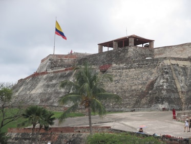 A historical fortress with large stone walls and a flat open area in the foreground. A palm tree is situated near the center, and several people can be seen walking around. A prominent flagpole bears the Colombian flag atop the fort. The sky is overcast, contributing to a subdued lighting quality.