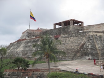 A historical fortress with large stone walls and a flat open area in the foreground. A palm tree is situated near the center, and several people can be seen walking around. A prominent flagpole bears the Colombian flag atop the fort. The sky is overcast, contributing to a subdued lighting quality.