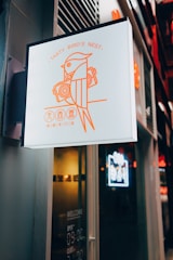 A sign featuring a stylized illustration of a bird holding a pair of speakers, accompanied by the text 'Tasty Bird's Nest' in orange. Below, there are Chinese characters. The sign is mounted on the exterior of a building, with a dark and modern design.