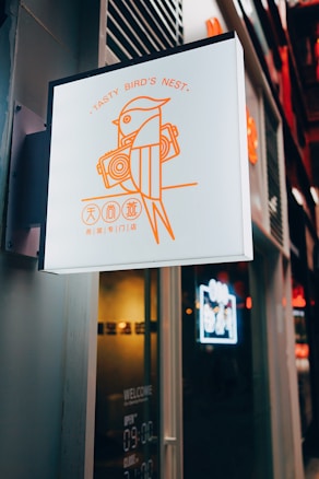 A sign featuring a stylized illustration of a bird holding a pair of speakers, accompanied by the text 'Tasty Bird's Nest' in orange. Below, there are Chinese characters. The sign is mounted on the exterior of a building, with a dark and modern design.
