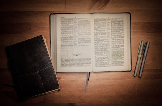 An open book with text is centered on a wooden surface, accompanied by a closed book on the left and three pens on the right.