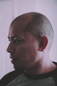 A side profile of a bald man with visible facial stubble, wearing a dark t-shirt. The lighting is soft and creates shadows on his face, giving a contemplative look.