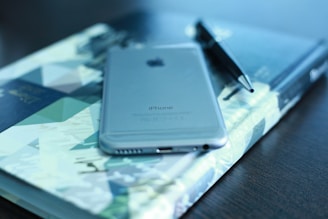 A sleek silver smartphone is placed on top of a colorful, geometric-patterned book. Beside it lies a black pen, all resting on a dark wooden table. The image conveys a sense of organization and modernity.
