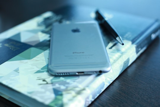 A sleek silver smartphone is placed on top of a colorful, geometric-patterned book. Beside it lies a black pen, all resting on a dark wooden table. The image conveys a sense of organization and modernity.