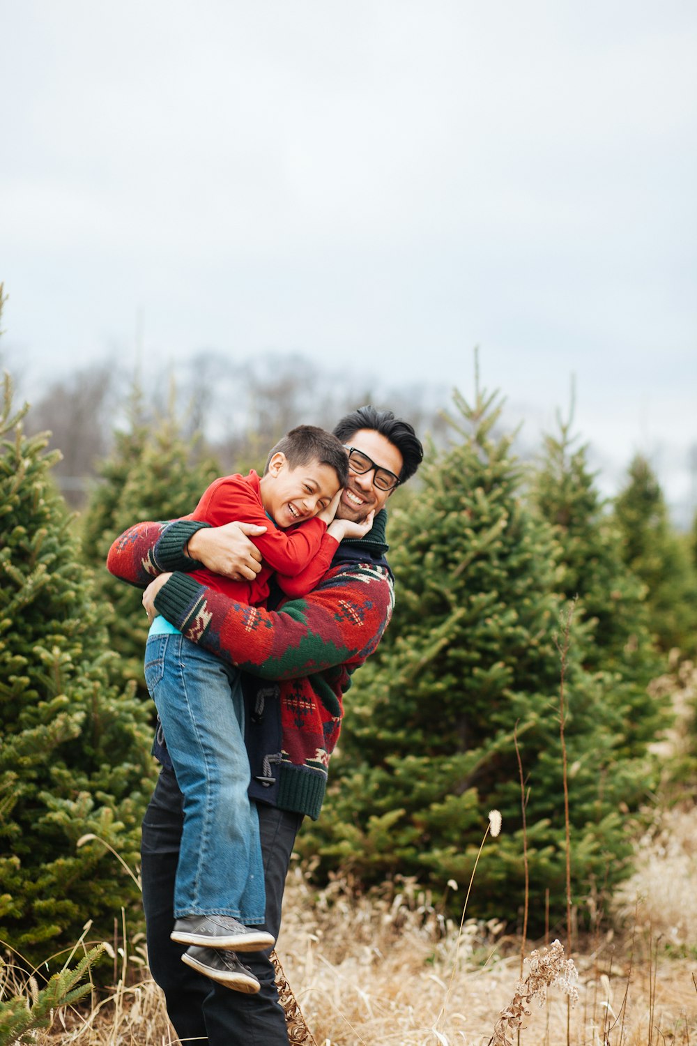 man carrying boy while standing and smiling near pine trees