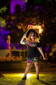 A person stands confidently wearing a dress decorated with colorful floral patterns, a large black hat, and makeup that mimics a traditional Dia de los Muertos skull design. The background features a dimly lit outdoor space with vibrant purple lighting and hanging decorations, creating a festive and atmospheric setting.
