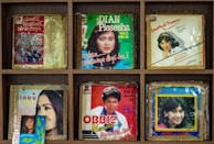 A collection of vintage album covers arranged in a wooden shelf. The covers feature portraits of various artists, with colorful designs and titles in different fonts. Each album is encased in plastic, showing signs of age and wear. The overall layout is organized and symmetrically arranged.