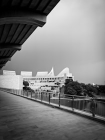 An architectural structure with modern design elements, featuring large angular roofs and intricate paneling, stands surrounded by greenery. The foreground includes a wide walkway with a railing, leading towards the building. The black-and-white tone adds a dramatic and timeless feel.