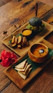 An elegantly arranged meal set on wooden trays atop a rustic table. One tray features a charcoal burger with fresh greens, accompanied by a small bowl of dipping sauce and thick-cut potato wedges. The second tray contains a bowl of soup with a garnish, and neatly folded flatbread pieces beside a vibrant red hibiscus flower.