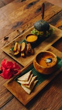 An elegantly arranged meal set on wooden trays atop a rustic table. One tray features a charcoal burger with fresh greens, accompanied by a small bowl of dipping sauce and thick-cut potato wedges. The second tray contains a bowl of soup with a garnish, and neatly folded flatbread pieces beside a vibrant red hibiscus flower.