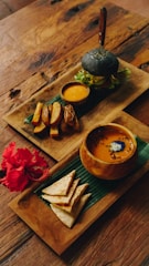 An elegantly arranged meal set on wooden trays atop a rustic table. One tray features a charcoal burger with fresh greens, accompanied by a small bowl of dipping sauce and thick-cut potato wedges. The second tray contains a bowl of soup with a garnish, and neatly folded flatbread pieces beside a vibrant red hibiscus flower.