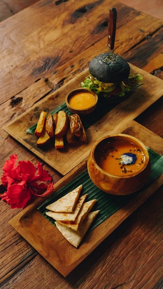 An elegantly arranged meal set on wooden trays atop a rustic table. One tray features a charcoal burger with fresh greens, accompanied by a small bowl of dipping sauce and thick-cut potato wedges. The second tray contains a bowl of soup with a garnish, and neatly folded flatbread pieces beside a vibrant red hibiscus flower.