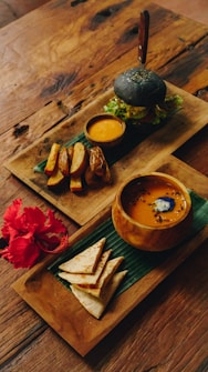 An elegantly arranged meal set on wooden trays atop a rustic table. One tray features a charcoal burger with fresh greens, accompanied by a small bowl of dipping sauce and thick-cut potato wedges. The second tray contains a bowl of soup with a garnish, and neatly folded flatbread pieces beside a vibrant red hibiscus flower.