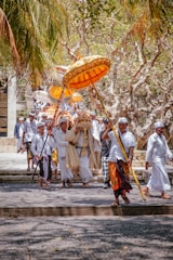 A traditional procession featuring people dressed in white attire, some carrying ornate umbrellas. The scene is set outdoors, under palm trees with dry, leafless branches, suggesting a cultural or religious event.