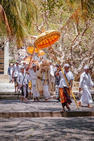 A traditional procession featuring people dressed in white attire, some carrying ornate umbrellas. The scene is set outdoors, under palm trees with dry, leafless branches, suggesting a cultural or religious event.