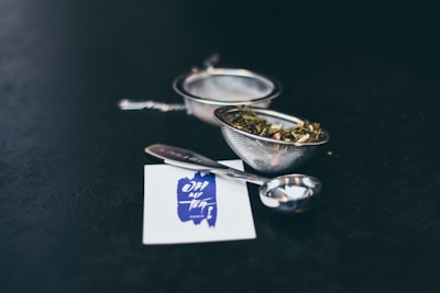 A set of metal tea strainers with leaves inside are placed on a dark surface alongside a measuring spoon and a business card featuring a blue design.