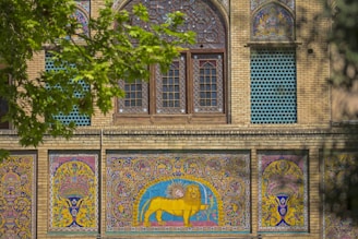 Intricate Persian-style mosaic tiles adorn the facade of a historical building. The design includes vibrant colors and detailed patterns with a prominent depiction of a lion and sun motif. Leafy green branches partially frame the scene.