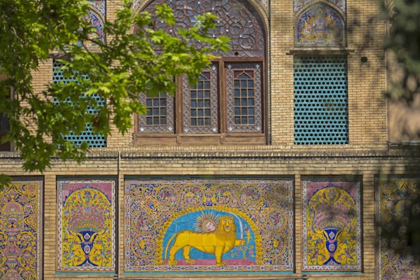 Intricate Persian-style mosaic tiles adorn the facade of a historical building. The design includes vibrant colors and detailed patterns with a prominent depiction of a lion and sun motif. Leafy green branches partially frame the scene.