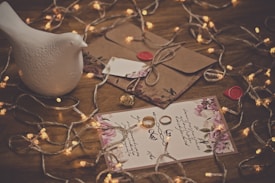A decorative white ceramic bird is positioned on a wooden surface surrounded by delicate string lights. Below, there are floral-themed envelopes sealed with red wax, and an ornate piece of jewelry similar in appearance to a ring. A card embellished with elegant script and floral patterns completes the arrangement.