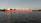 A large red cargo ship is sailing on a calm body of water with trees and a faint outline of structures in the background.