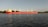 A large red cargo ship is sailing on a calm body of water with trees and a faint outline of structures in the background.