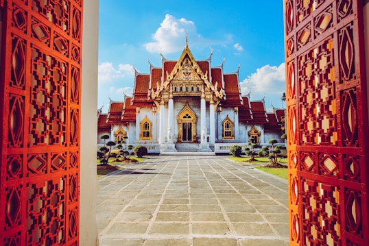 A stunning ornate temple with a golden roof is framed between two elaborately carved red doors. The temple, with its intricate architecture, sits centrally under a vibrant blue sky scattered with white clouds. The entrance path is made of large stone tiles, flanked by neatly trimmed shrubs on either side.
