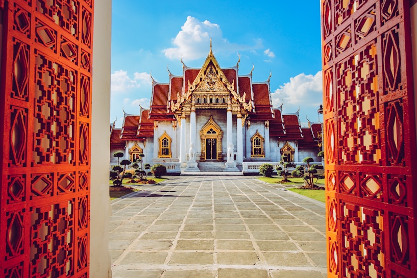 A stunning ornate temple with a golden roof is framed between two elaborately carved red doors. The temple, with its intricate architecture, sits centrally under a vibrant blue sky scattered with white clouds. The entrance path is made of large stone tiles, flanked by neatly trimmed shrubs on either side.