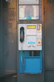 A vintage public payphone with a handset, keypad, and coin slot is mounted on an old, weathered metal panel. The surrounding metal surface shows signs of rust and graffiti marks, adding to the aged appearance.
