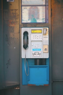 A vintage public payphone with a handset, keypad, and coin slot is mounted on an old, weathered metal panel. The surrounding metal surface shows signs of rust and graffiti marks, adding to the aged appearance.