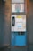 A vintage public payphone with a handset, keypad, and coin slot is mounted on an old, weathered metal panel. The surrounding metal surface shows signs of rust and graffiti marks, adding to the aged appearance.