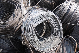 Large coils of metallic wire are tightly wound and layered together. The smooth, shiny surface of the wires reflects light prominently, creating a complex texture of curved lines. The wire is neatly stacked, giving the impression of organized industrial material ready for use.