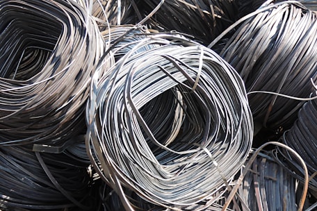 Large coils of metallic wire are tightly wound and layered together. The smooth, shiny surface of the wires reflects light prominently, creating a complex texture of curved lines. The wire is neatly stacked, giving the impression of organized industrial material ready for use.