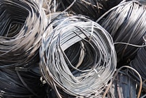 Large coils of metallic wire are tightly wound and layered together. The smooth, shiny surface of the wires reflects light prominently, creating a complex texture of curved lines. The wire is neatly stacked, giving the impression of organized industrial material ready for use.